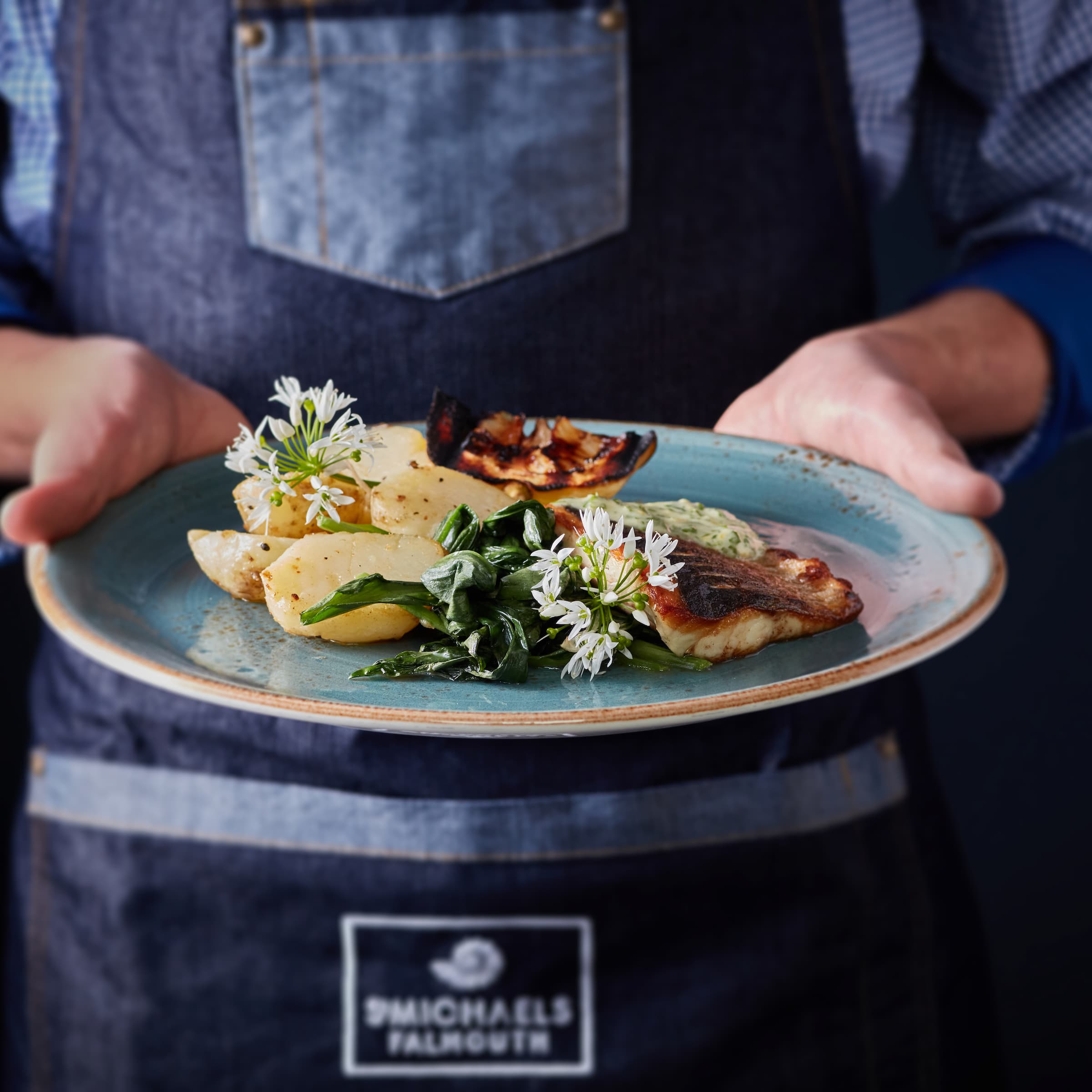 Server in a dark apron presents a colourful gourmet plate with grilled fish, vegetables, herbs, and white edible flowers, invitingly styled.