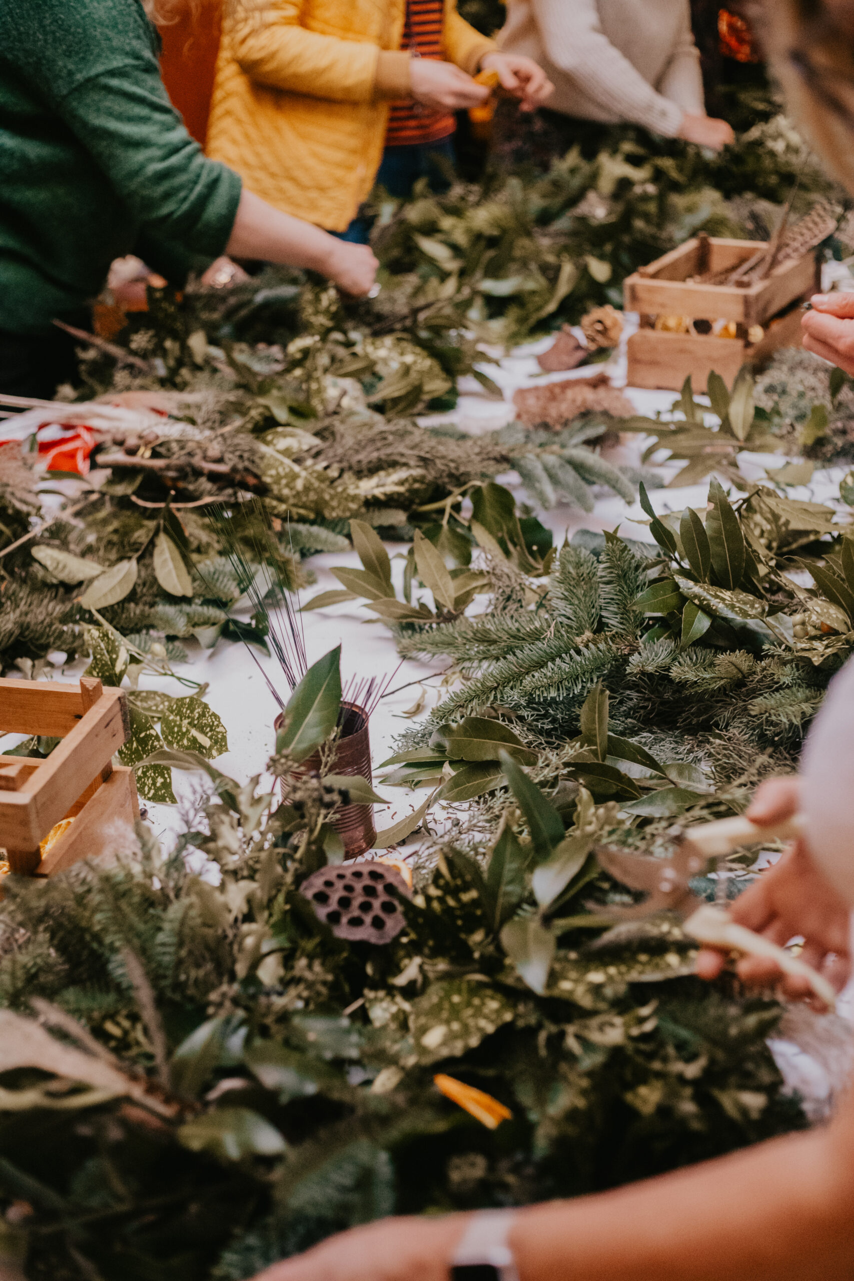 Guests making wreaths around a long table covered in fresh winter foliage and wooden trays of decorations